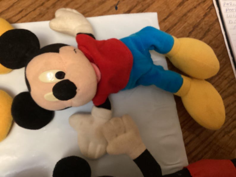 Puede incluir: Un peluche de Mickey Mouse, con una camisa roja, pantalones cortos azules, zapatos amarillos y guantes blancos. El ic&oacute;nico personaje tiene orejas negras y una cara bronceada con ojos grandes y redondos. El juguete est&aacute; sobre una superficie blanca.