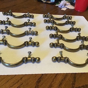May include: Twelve antique brass cabinet door pulls with a rounded, arched design. Each pull has a decorative, fluted, cylindrical shape with a rounded knob at each end.