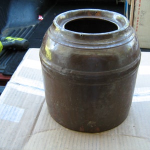 May include: A brown ceramic crock with a wide opening and a slightly textured surface. The crock has a simple design with a few raised rings around the body.