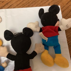 Puede incluir: Varios peluches de Mickey Mouse est&aacute;n dispuestos sobre una superficie blanca. Los peluches tienen orejas negras, guantes blancos, camisas rojas, pantalones azules y zapatos amarillos. Un juguete est&aacute; mirando al espectador, con los brazos extendidos.