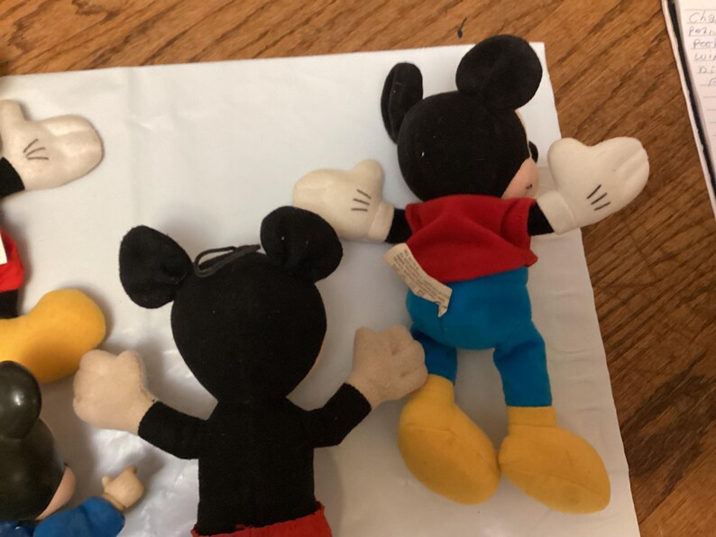 Puede incluir: Varios peluches de Mickey Mouse est&aacute;n dispuestos sobre una superficie blanca. Los peluches tienen orejas negras, guantes blancos, camisas rojas, pantalones azules y zapatos amarillos. Un juguete est&aacute; mirando al espectador, con los brazos extendidos.