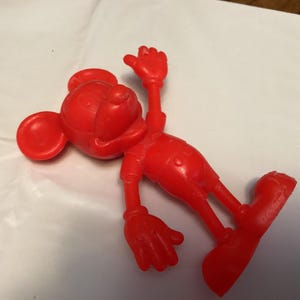 May include: A bright red Mickey Mouse figurine in a playful pose. The figure is lying on its side with one arm raised and the other extended. The classic cartoon character is wearing shorts and shoes.