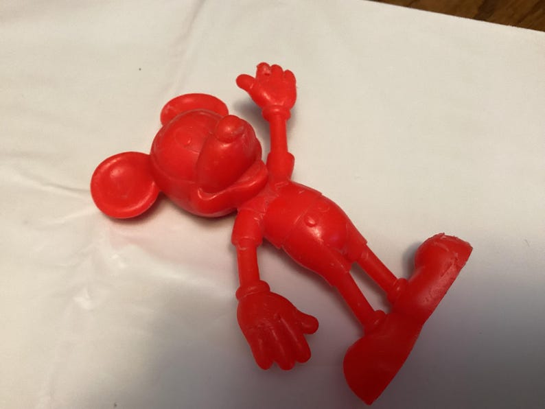 May include: A bright red Mickey Mouse figurine in a playful pose. The figure is lying on its side with one arm raised and the other extended. The classic cartoon character is wearing shorts and shoes.
