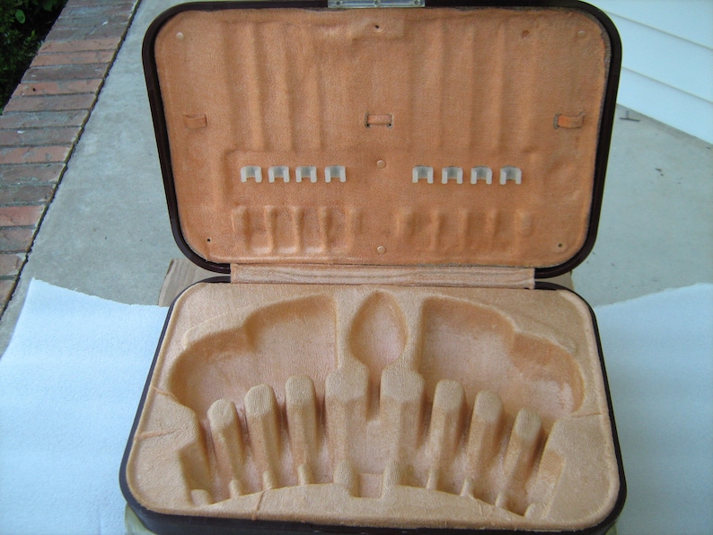 May include: A brown leatherette case with a beige velvet interior. The case has a hinged lid and is designed to hold cutlery. The interior has slots for forks, knives, and spoons.