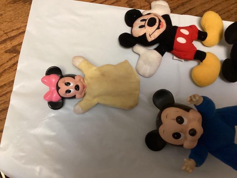 Puede incluir: Una colecci&oacute;n de peluches de Mickey Mouse y Minnie Mouse. Mickey Mouse viste su atuendo cl&aacute;sico con pantalones cortos rojos, orejas negras y zapatos amarillos. Minnie Mouse tiene un lazo rosa. Un Mickey Mouse lleva un traje azul.