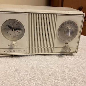 May include: A vintage General Electric clock radio in off-white. The radio features a clock face on the left, a speaker grill in the center, and a tuning dial on the right. The word "VOLUME" is printed below the dial.