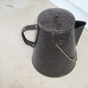 May include: Black enamel camping coffee pot with a speckled finish and a lid. The pot has a handle and a wire bail.
