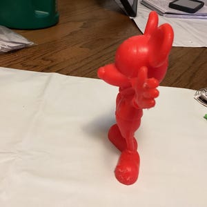 May include: A solid red figurine of a cartoon character, posed with one arm raised and the other extended. The figure has large ears and a smiling expression. The figurine is standing on a white surface.
