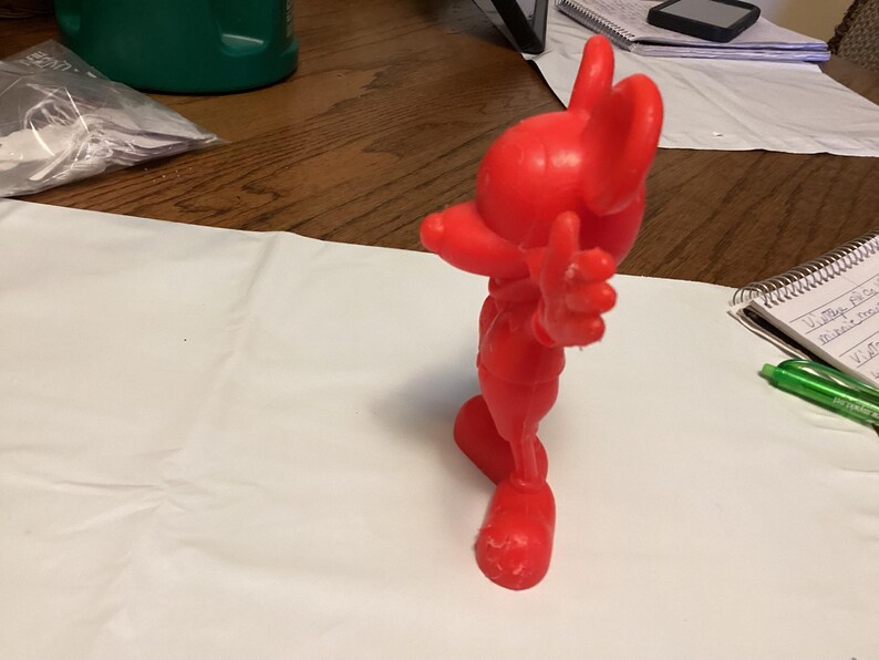 May include: A solid red figurine of a cartoon character, posed with one arm raised and the other extended. The figure has large ears and a smiling expression. The figurine is standing on a white surface.