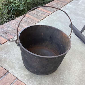 May include: A round, black cast iron pot with a deep basin and a curved metal handle attached to the sides. The interior shows signs of oxidation and wear, with a dark, weathered surface texture throughout the exterior and rim.