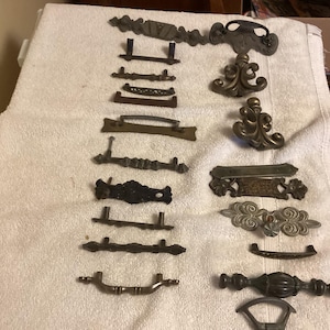 May include: A collection of antique drawer pulls and handles in various styles and finishes, including brass, bronze, and black metal. The hardware features ornate designs and classic shapes, perfect for vintage furniture restoration.