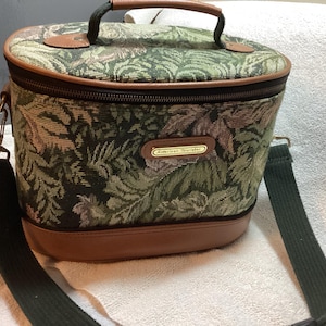 May include: An oval-shaped American Tourister travel case featuring a green and brown botanical tapestry pattern. The bag includes a brown leather-like base, a top handle, a zippered closure, and a detachable dark green fabric shoulder strap.