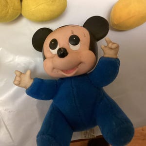 Puede incluir: Mu&ntilde;eco vintage de Mickey Mouse con un mono azul. El mu&ntilde;eco tiene orejas negras, boca rosa y ojos negros. Los brazos del mu&ntilde;eco est&aacute;n levantados y parece estar hecho de un material suave. Otros art&iacute;culos de felpa amarillos son visibles en el fondo.