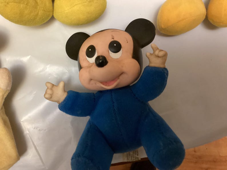 Puede incluir: Mu&ntilde;eco vintage de Mickey Mouse con un mono azul. El mu&ntilde;eco tiene orejas negras, boca rosa y ojos negros. Los brazos del mu&ntilde;eco est&aacute;n levantados y parece estar hecho de un material suave. Otros art&iacute;culos de felpa amarillos son visibles en el fondo.