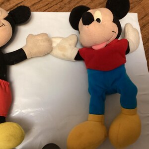Puede incluir: Dos peluches de Mickey Mouse. Un Mickey lleva pantalones cortos rojos con botones blancos, una camisa negra y zapatos amarillos. El otro Mickey lleva pantalones azules, una camisa roja y zapatos amarillos. Ambos tienen orejas negras y guantes blancos.
