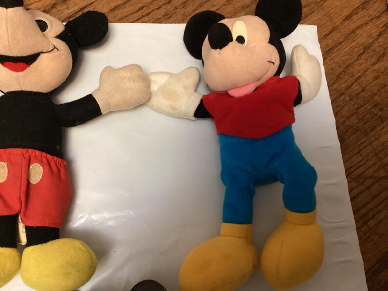 Puede incluir: Dos peluches de Mickey Mouse. Un Mickey lleva pantalones cortos rojos con botones blancos, una camisa negra y zapatos amarillos. El otro Mickey lleva pantalones azules, una camisa roja y zapatos amarillos. Ambos tienen orejas negras y guantes blancos.