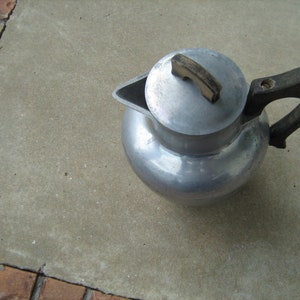 May include: A silver metal teapot with a black handle and a lid. The teapot is sitting on a gray concrete surface.