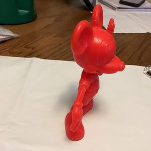 May include: A bright red, vintage-style figurine of a cartoon mouse. The figure has large ears, a long snout, and is posed in a walking stance. The toy is made of a solid material and stands on a white surface.