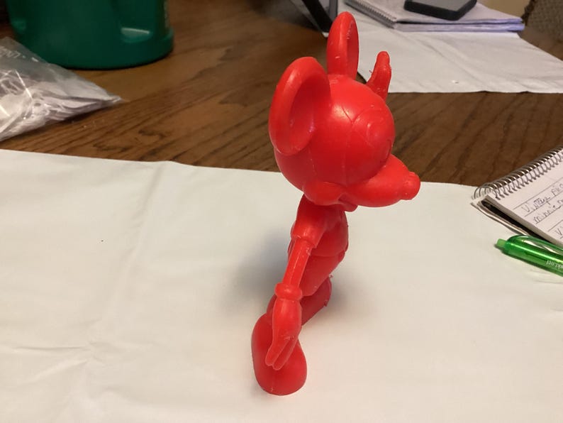 May include: A bright red, vintage-style figurine of a cartoon mouse. The figure has large ears, a long snout, and is posed in a walking stance. The toy is made of a solid material and stands on a white surface.