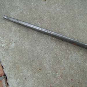 May include: A long, thin, silver metal rod with a rounded end. The rod is lying on a gray concrete surface.