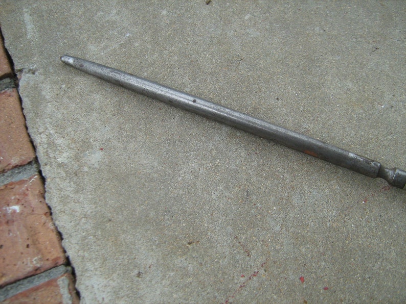 May include: A long, thin, silver metal rod with a rounded end. The rod is lying on a gray concrete surface.