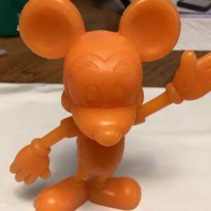 May include: An orange plastic figurine of a cartoon mouse with large ears, a long nose, and gloved hands. The mouse is posed with one arm raised in a waving gesture. The figurine is standing on a white surface.