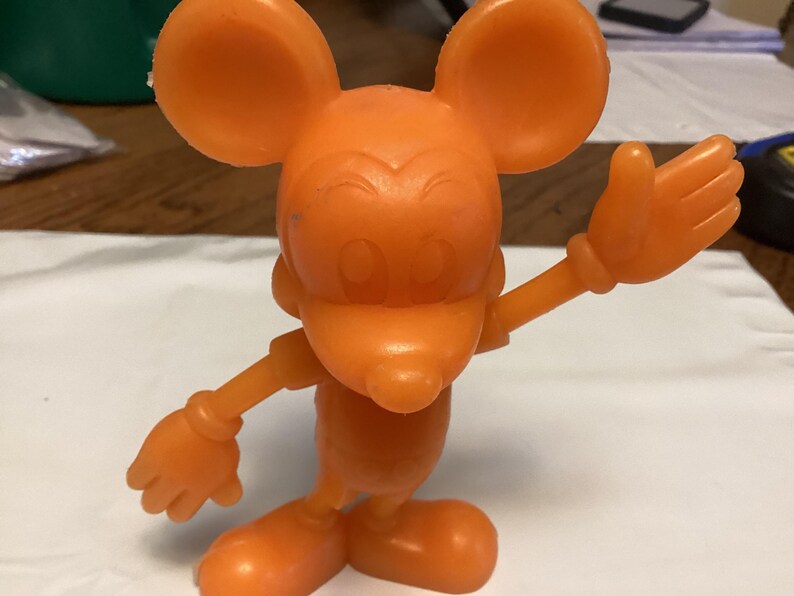 May include: An orange plastic figurine of a cartoon mouse with large ears, a long nose, and gloved hands. The mouse is posed with one arm raised in a waving gesture. The figurine is standing on a white surface.