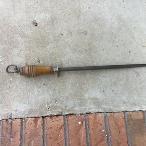 May include: A knife sharpening steel with a wooden handle and a metal sharpening rod. The handle is light brown and has a metal ring at the end. The sharpening rod is dark gray and tapers to a point. Kitchen tool.