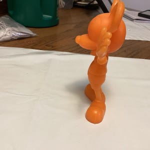 May include: An orange plastic figurine of a stylized character in a dynamic pose. The figure has large, rounded features, including a head, ears, and feet. The figurine is standing on a white surface.