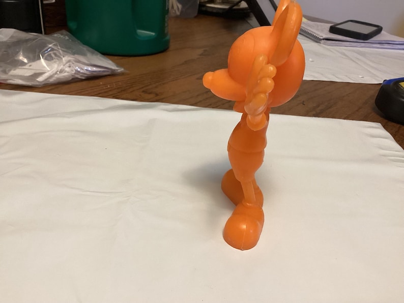 May include: An orange plastic figurine of a stylized character in a dynamic pose. The figure has large, rounded features, including a head, ears, and feet. The figurine is standing on a white surface.