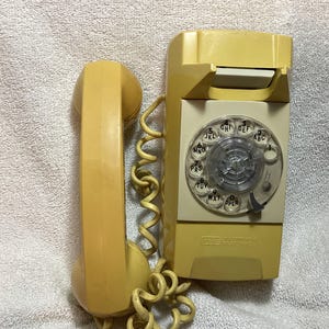 May include: A vintage wall-mounted rotary telephone in a pale yellow hue. The device features a circular clear plastic dial with black numbers and letters, a matching yellow handset, and a coiled cord connecting the handset to the main base unit.