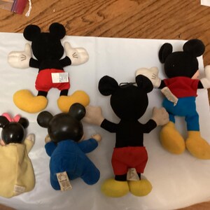 Puede incluir: Una colecci&oacute;n de peluches de Mickey Mouse y Minnie Mouse. Los juguetes de Mickey Mouse tienen cuerpos negros, pantalones cortos rojos, zapatos amarillos y guantes blancos. Minnie Mouse tiene un lazo rosa y un vestido amarillo. Los juguetes est&aacute;n sobre una superficie blanca.
