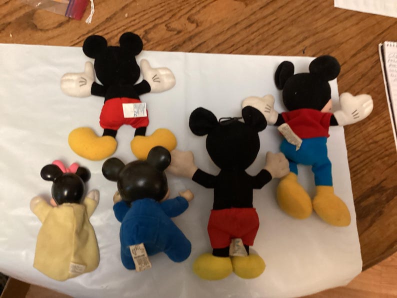Puede incluir: Una colecci&oacute;n de peluches de Mickey Mouse y Minnie Mouse. Los juguetes de Mickey Mouse tienen cuerpos negros, pantalones cortos rojos, zapatos amarillos y guantes blancos. Minnie Mouse tiene un lazo rosa y un vestido amarillo. Los juguetes est&aacute;n sobre una superficie blanca.