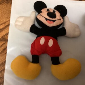 Puede incluir: Un peluche de Mickey Mouse con cabeza y orejas negras, torso negro, pantalones cortos rojos con botones blancos, guantes blancos y zapatos amarillos. El juguete sonr&iacute;e, con la lengua rosa visible.