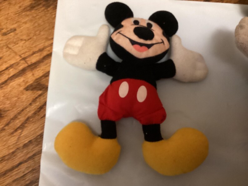 Puede incluir: Un peluche de Mickey Mouse con cabeza y orejas negras, torso negro, pantalones cortos rojos con botones blancos, guantes blancos y zapatos amarillos. El juguete sonr&iacute;e, con la lengua rosa visible.