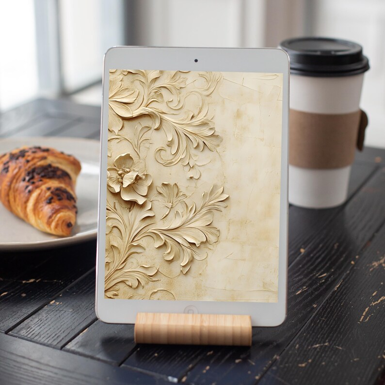 May include: A tablet displaying a digital wallpaper with an intricate carved floral design in shades of beige and cream. The tablet is propped up on a wooden stand.