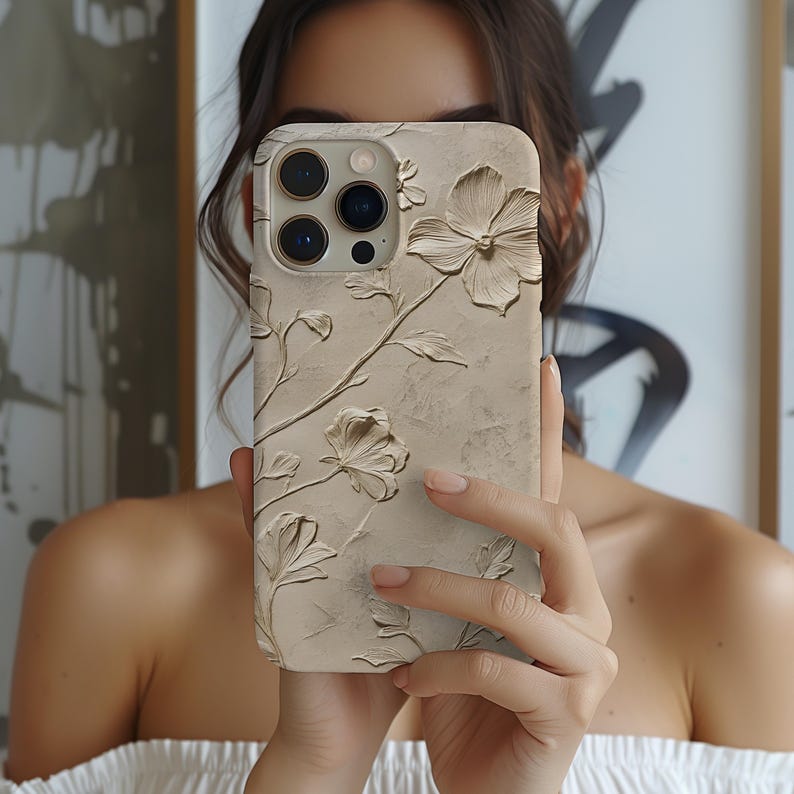 May include: A beige phone case with a raised floral design. The case has a textured surface and features a three-lens camera cutout.
