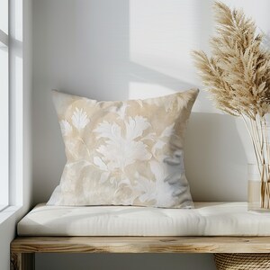 May include: A beige throw pillow with a white floral pattern. The pillow is sitting on a light brown wooden bench with a white cushion.