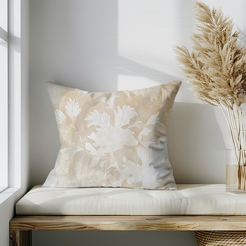May include: A beige throw pillow with a white floral pattern. The pillow is sitting on a light brown wooden bench with a white cushion.