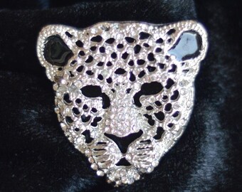 May include: A silver-toned leopard head brooch with black enamel accents and clear crystal embellishments. The brooch is set against a backdrop of black velvet fabric, creating a striking contrast. The brooch is a fashion accessory.