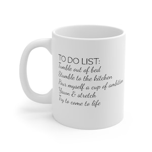 May include: A white ceramic mug featuring black printed text that reads: TO DO LIST: Tumble out of bed, Stumble to the kitchen, Pour myself a cup of ambition, Yawn & stretch, Try to come to life. The mug has a curved handle on the left side.