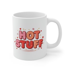 Puede incluir: Taza de cerámica blanca con la frase "HOT STUFF" en fuente de burbujas. El texto es en tonos rojos y naranjas, con corazones y estrellas decorativas. La taza tiene un asa curva.