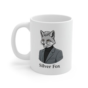 May include: White ceramic mug featuring a cartoon illustration of a silver fox wearing a gray blazer and black turtleneck. The text "Silver Fox" is printed below the illustration. The mug has a curved handle.