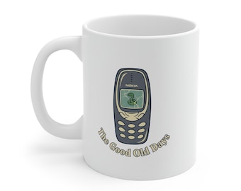 Może przedstawiać: Biały ceramiczny kubek z grafiką retro telefonu Nokia. Telefon jest szary i zielony, z grą w węża na ekranie. Napis "The Good Old Days" jest w okrągłym wzorze wokół telefonu.