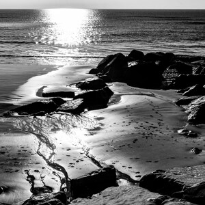 Puede incluir: Una fotografía en blanco y negro de una playa rocosa con el sol reflejándose en el agua. El agua retrocede, dejando un patrón de ondas en la arena.
