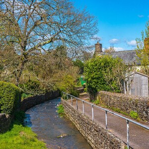 May include: A stone path leads to a small village with a stone wall and a stream running alongside it. The stream is surrounded by lush green grass and trees.