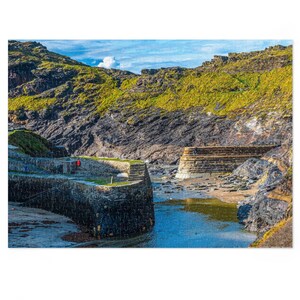 Pode incluir: Um quebra-cabeça completo retratando uma cena costeira. A imagem mostra um porto de pedra com um mar azul, falésias rochosas e vegetação verde sob um céu parcialmente nublado. O quebra-cabeça está completo e pronto para ser exibido.