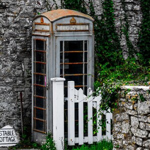May include: A vintage, gray, public telephone booth with a white picket fence in front of it. The booth is set against a stone wall covered in green ivy.