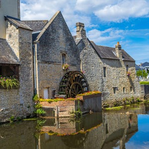 May include: A stone building with a water wheel in front of it. The building is old and has a rustic charm. The water wheel is made of wood and is turning in the water. The building is surrounded by trees and flowers.