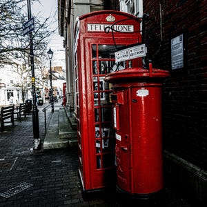 Pode incluir: Uma cabine telefônica vermelha e uma caixa de correio vermelha ficam lado a lado em uma rua de paralelepípedos. A cabine telefônica tem uma placa que diz "Telefone" e a caixa de correio tem uma placa que diz "Correios".